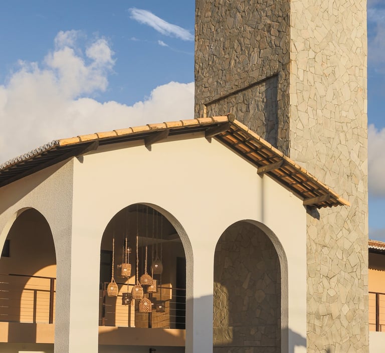 Modern villa with white arches, stone tower, and hanging wicker lanterns against a blue sky.