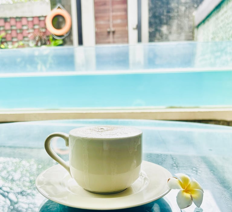 A white cappuccino cup on a glass table by a luxury swimming pool at a tropical villa resort.