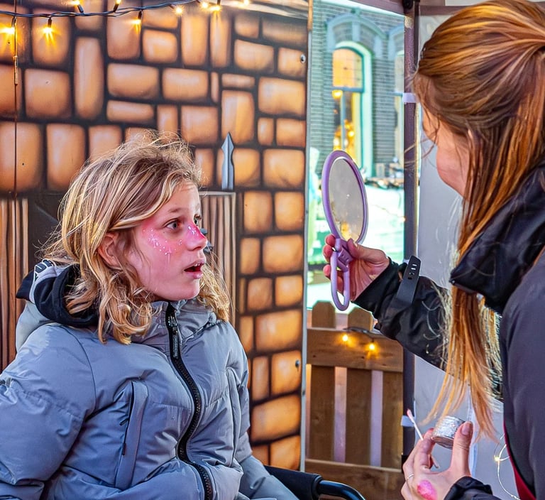 Een kind met roze gezichtsschmink kijkt met een verbaasde blik in een handspiegel die wordt vastgeho