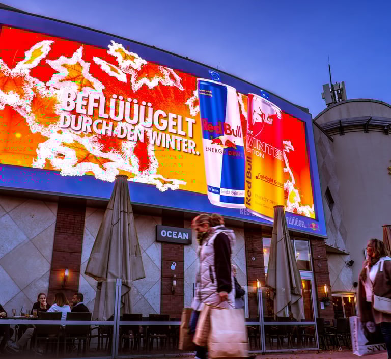 Large digital billboard advertising Red Bull Winter Edition above a crowded outdoor cafe at dusk.