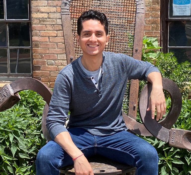 Hispanic man seated in a metal chair sculpture with a brick building and bushes close in the background