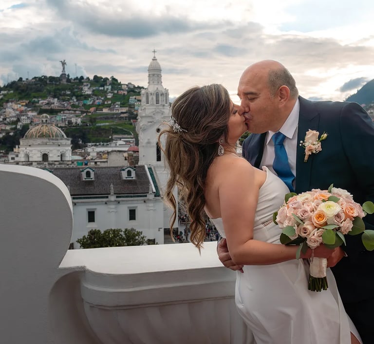 El novio besando a la novia mientras la agarra de la cintura en un terraza con vista al Panecillo en el centro histórico