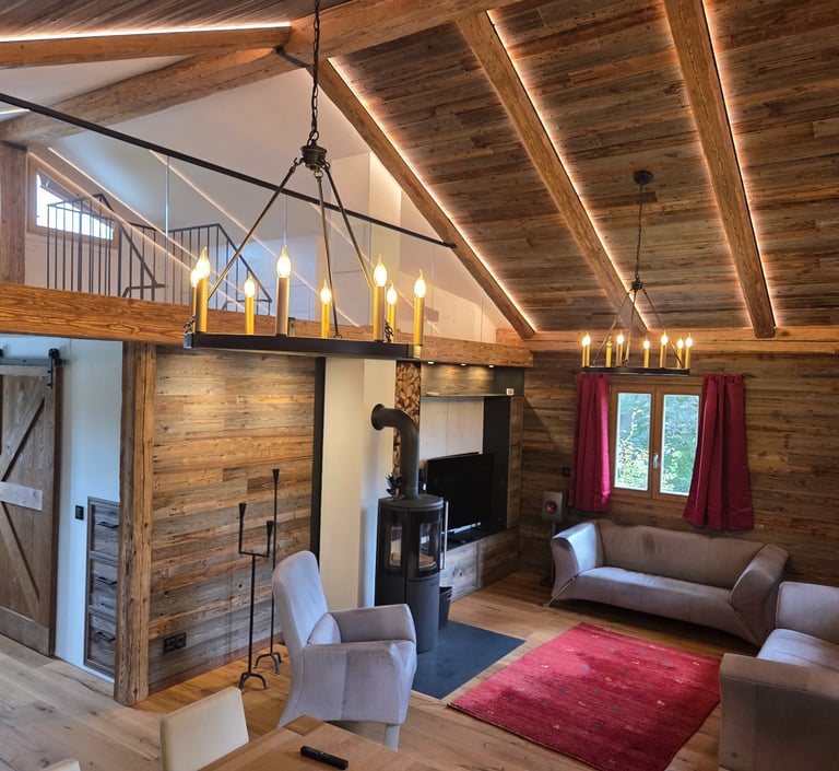 Großzügiges Wohnzimmer mit offenem Altholz-Dachstuhl, Galerie mit Glasgeländer, zwei Kronleuchtern und Kaminofen.