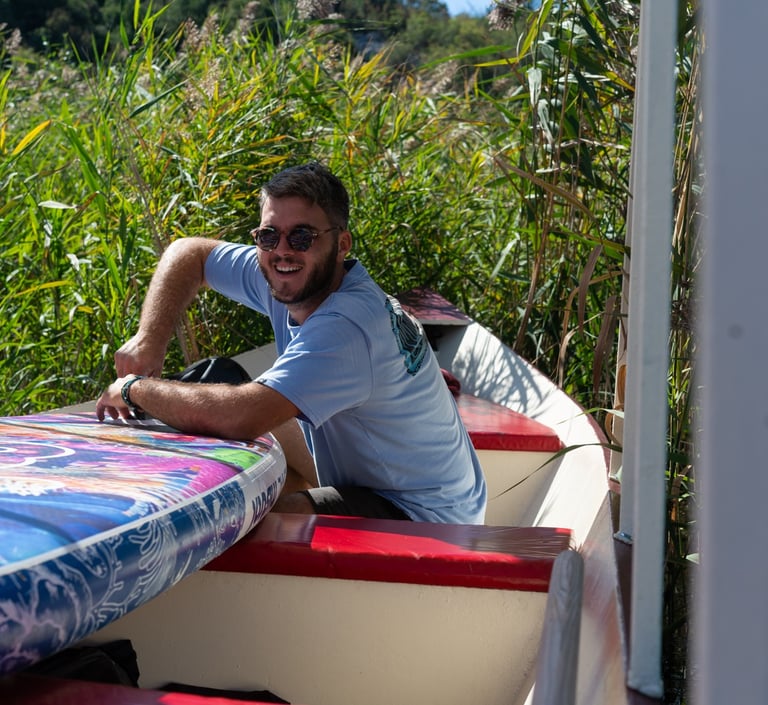 A smiling man with sunglasses sits in a boat with a colorful paddleboard near tall reeds.