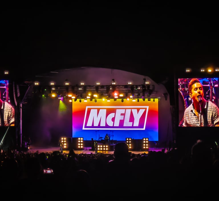McFly performing live on a brightly lit concert stage with large LED screens for fans at a night music festival.