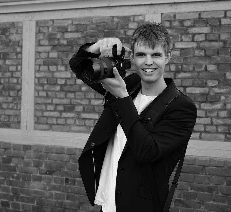 Schwarz-Weiß-Aufnahme von Joel Pingel mit Kamera vor einer Ziegelwand. Eventfotografie: Just Perspectives.