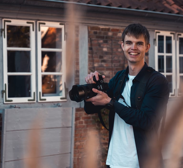 Lächelnder Hochzeitsfotograf Joel Pingel mit Kamera vor Backsteingebäude. Portrait: Just Perspectives.