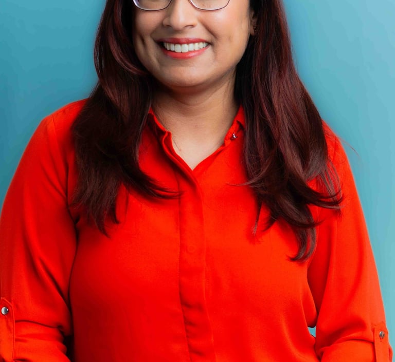A smiling woman in glasses stands against a blue background, wearing a vibrant red shirt and a friendly demeanor.