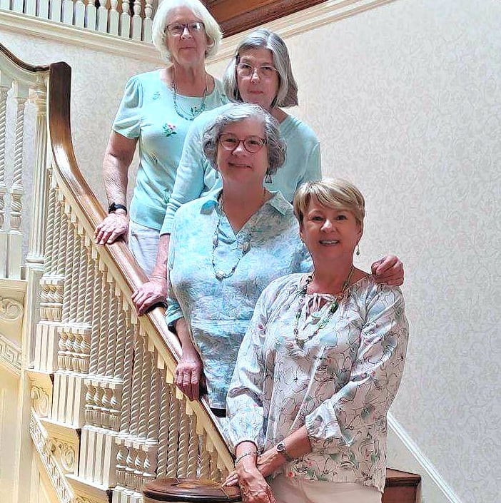 Four smiling senior women posing together on a historic wooden staircase in a bright home.