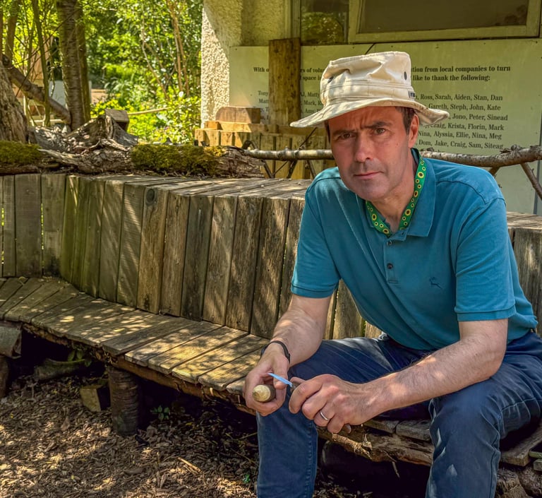 Simon is sitting on a wooden bench in the shade at Westbury Wildlife Park, whittling a mushroom.