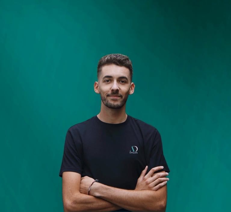 A young man in a black AD Cycling t-shirt posing with crossed arms against a teal background.
