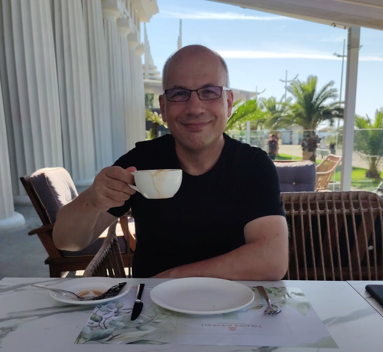 Smiling Heinz Roelfs in glasses enjoying coffee at an outdoor seaside terrace restaurant.