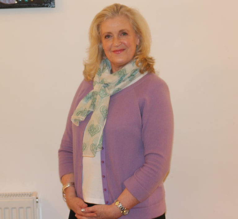 blonde haired woman smiling wearing light purple cardigan and green scarf