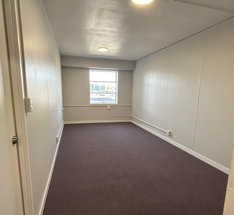 Small unfurnished office with dark carpet and 1 window on the far wall