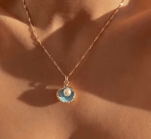 A simple gold necklace with half a shell as a pendant. A pearl hangs from the chain to sit in it.