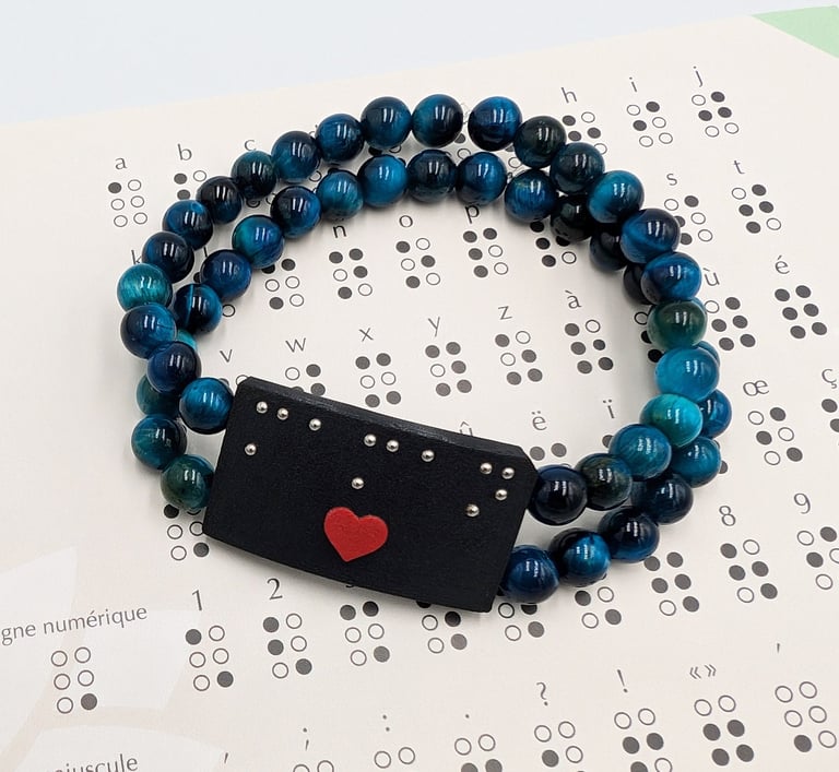 Bracelet perle bleue avec une plaque en braille et un cœur rouge, posé sur un alphabet braille.