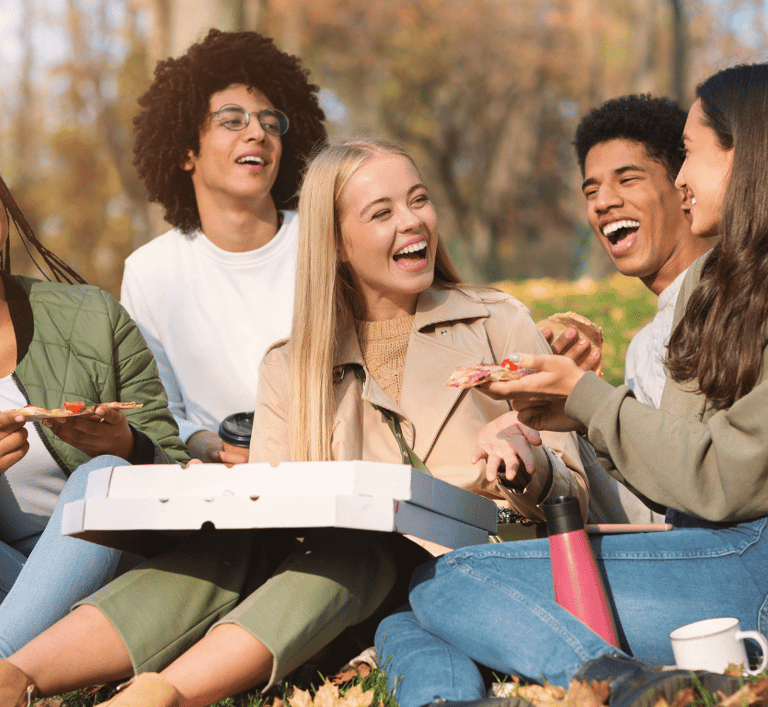 Diverse group of smiling friends enjoying a pizza picnic outdoors in a sunny autumn park.