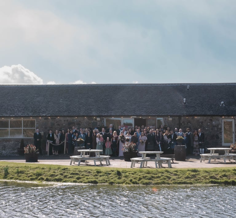 Wedding guests posing for a group photo outside a rustic stone barn venue by a scenic pond.