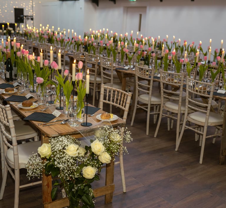 Wedding reception long wooden tables decorated with pink tulips, white roses, and lit candles.