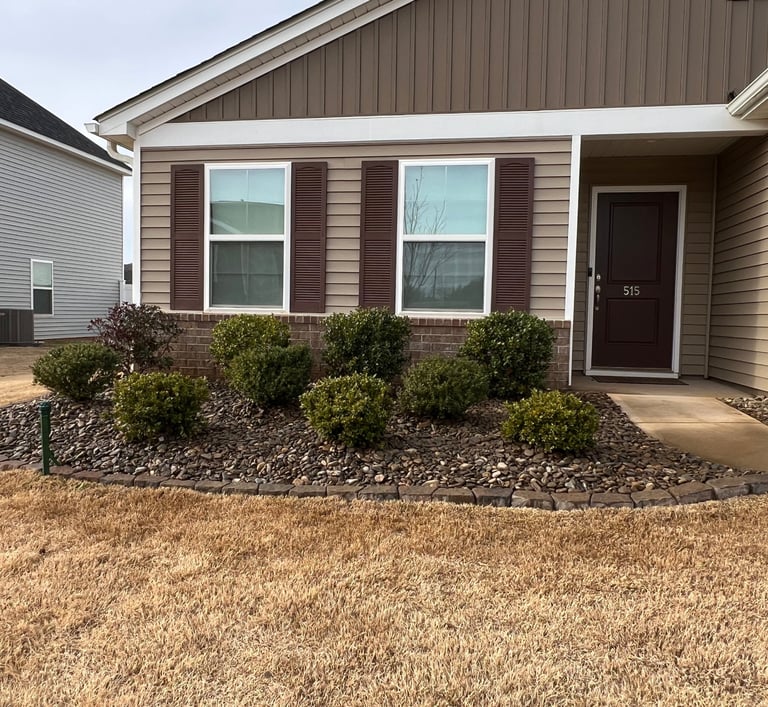 Flower bed in front of brown house with evergreen shrubs, cane rock for mulch and brown stone border.