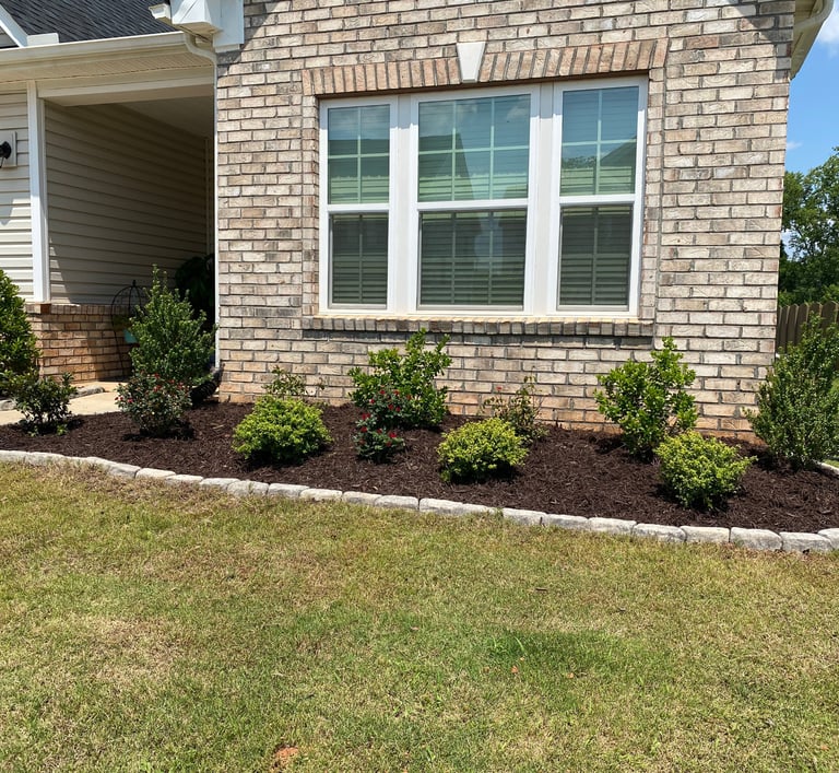 Flower bed in front of brick house with evergreen shrubs, brown mulch, and gray stone border.