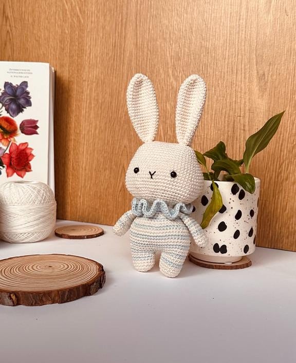 a small handmade amigurumi white bunny, with body in stripes, with a potted plant in the background