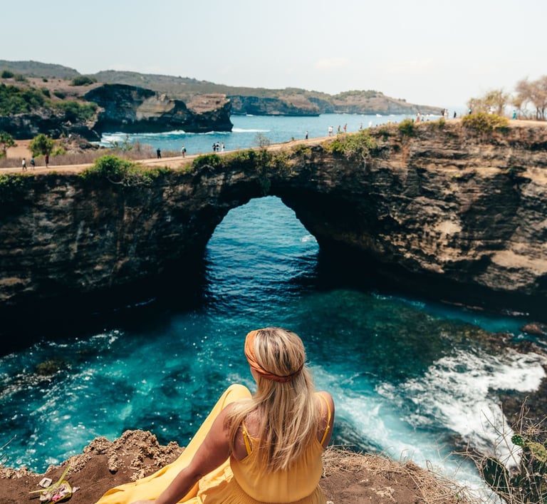 A blonde woman in a yellow dress sits overlooking Broken Beach in Nusa Penida, Bali.