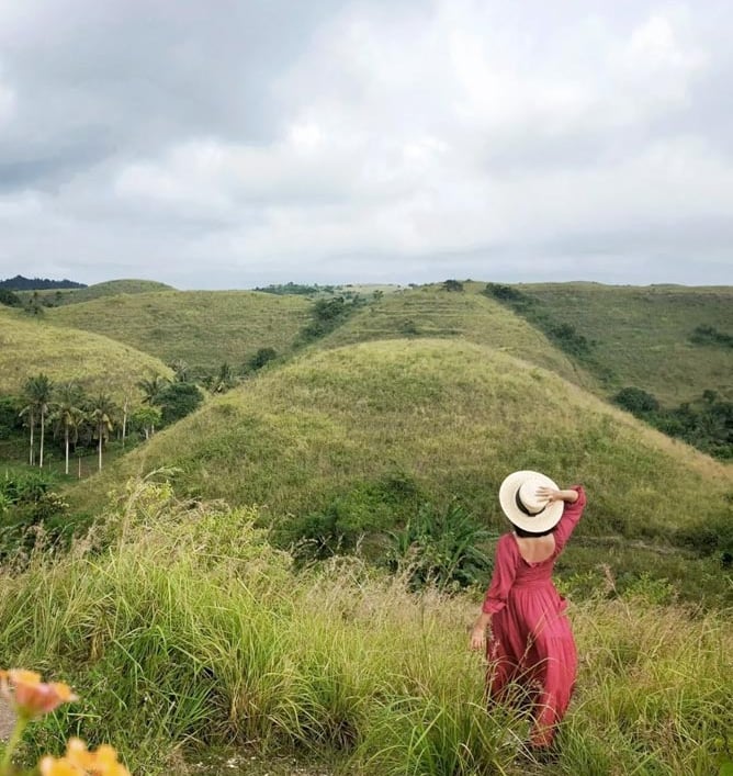 A woman in a red dress and sun hat overlooks lush green rolling hills and tropical landscape.