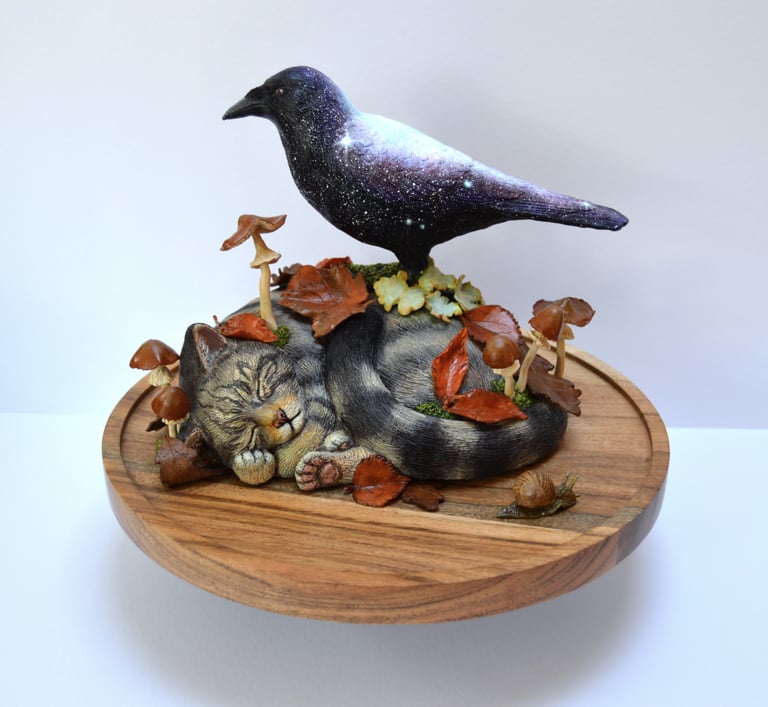 Handcrafted sculpture of a celestial galaxy crow perched on a sleeping tabby cat figurine with autumn leaves and mushrooms.