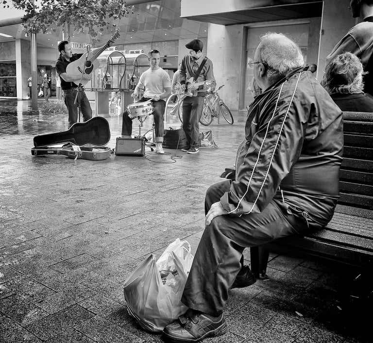 Older man sitting on mall bench listening to busking rock and roll group