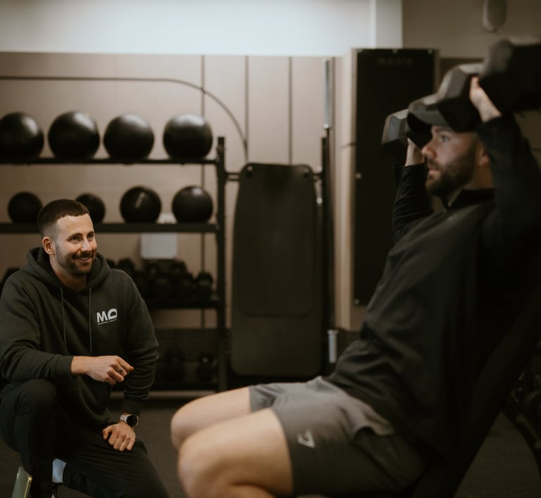 Trainer observing client’s seated dumbbell shoulder press in Leamington Spa gym, highlighting coaching and strength training.