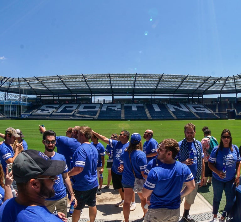 Crowd of fans in blue shirts socializing on the field at Sporting Kansas City stadium under a bright sky.