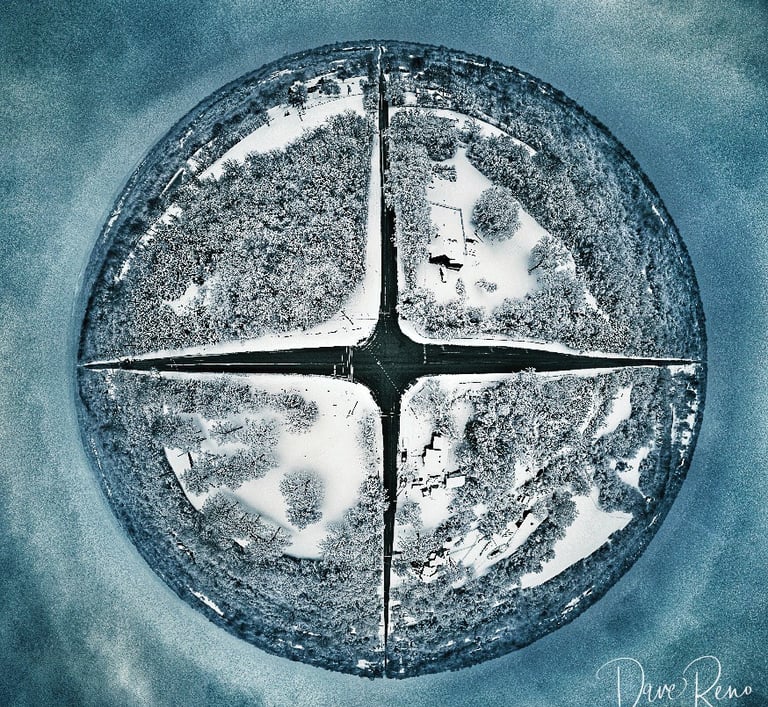 Tiny planet aerial view of a four-way street intersection surrounded by snow-covered trees and homes.