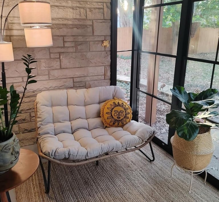 Cozy reading nook with a tufted papasan loveseat, sun pillow, and potted plants by a large window.