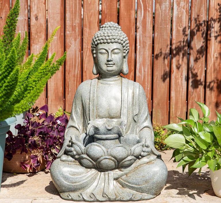 A stone Buddha garden statue sitting in a lotus pose near green plants and a wooden fence.
