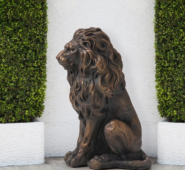 Majestic bronze-finished lion garden statue sitting between green hedges against a white wall.