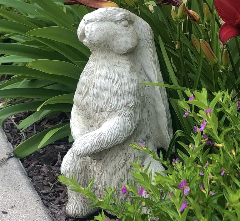 A decorative stone rabbit statue sitting in a garden among green leaves and purple flowers.