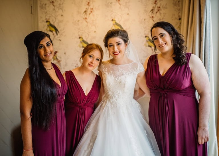 Smiling bride in a lace wedding dress posing with bridesmaids in plum purple dresses.
