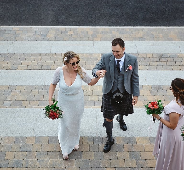 A smiling bride in a white dress and groom in a traditional Scottish kilt walk up stone stairs.