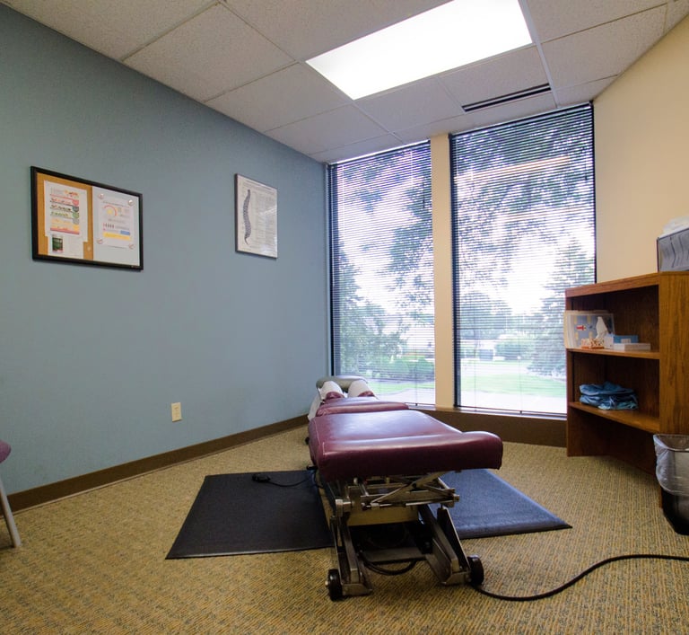 Clean chiropractic exam room with a treatment table, a bookcase of supplies, and a single chair.