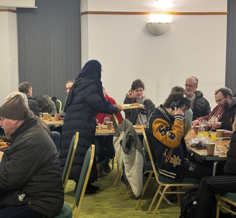 Diverse group of people gathering for a community meal event indoors at long dining tables.