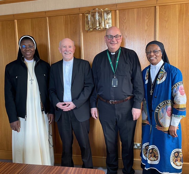 Four religious figures, including nuns and priests, stand together in a formal room with wood paneling, smiling warmly.