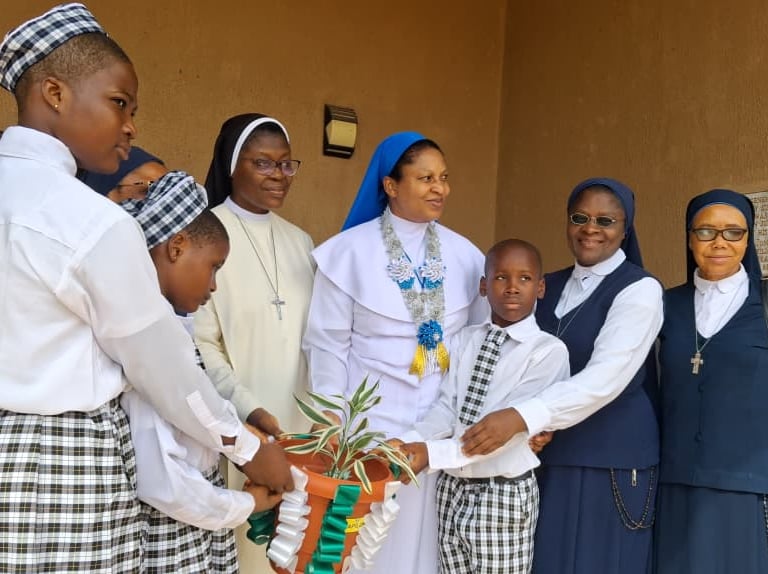 Catholic nuns and school children holding a potted plant together at a religious ceremony.
