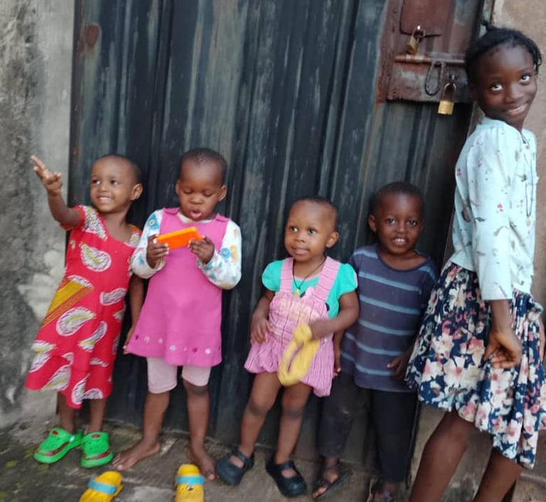Five happy young African children posing together in front of a dark metal door outdoors.
