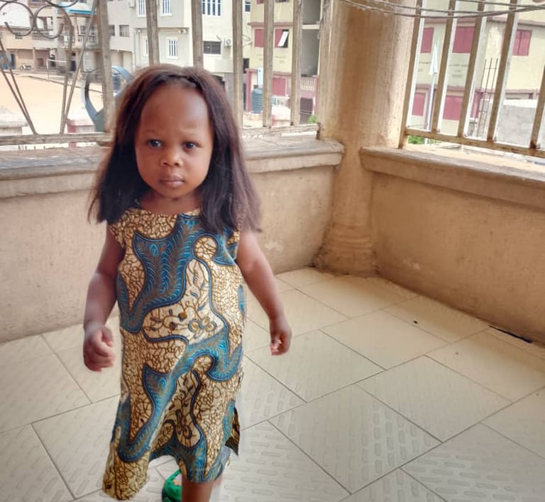 A young girl wearing a blue and yellow patterned Ankara dress walks on a tiled balcony.