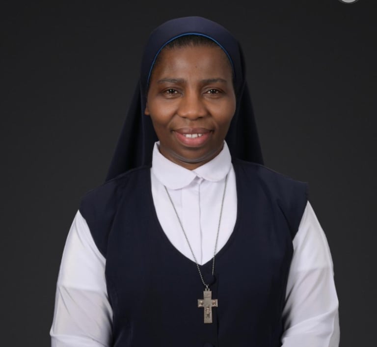 A smiling Catholic nun wearing a blue and white habit with a silver crucifix necklace against a dark background.