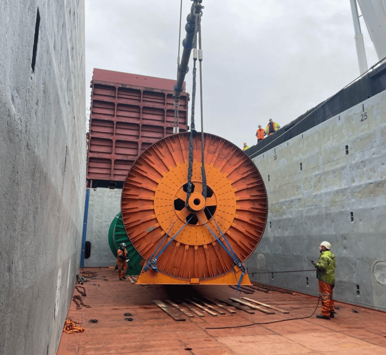 Industrial reel being lifted onto a cargo ship with workers and cranes