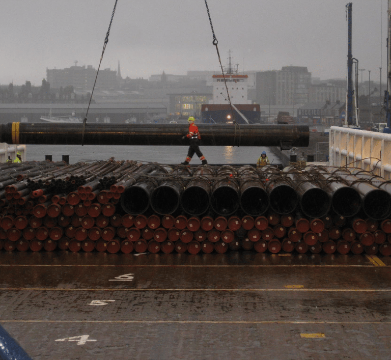 Worker guiding large pipe being unloaded from cargo ship with stacked pipes on deck
