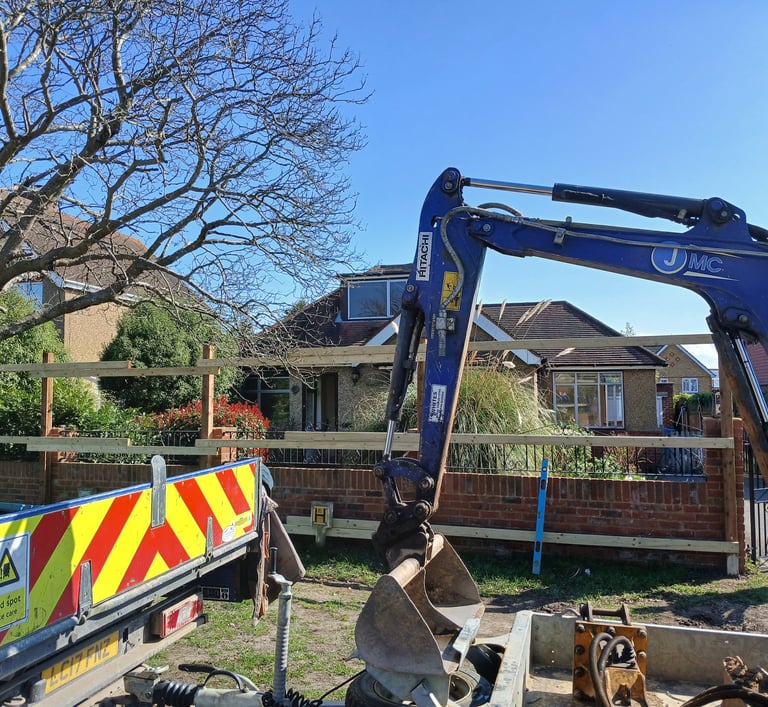 Blue Hitachi mini excavator and flatbed truck at a residential construction site near a wooden fence.