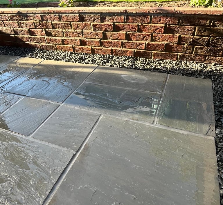 Wet grey flagstone patio tiles with black gravel borders and a red brick retaining wall.